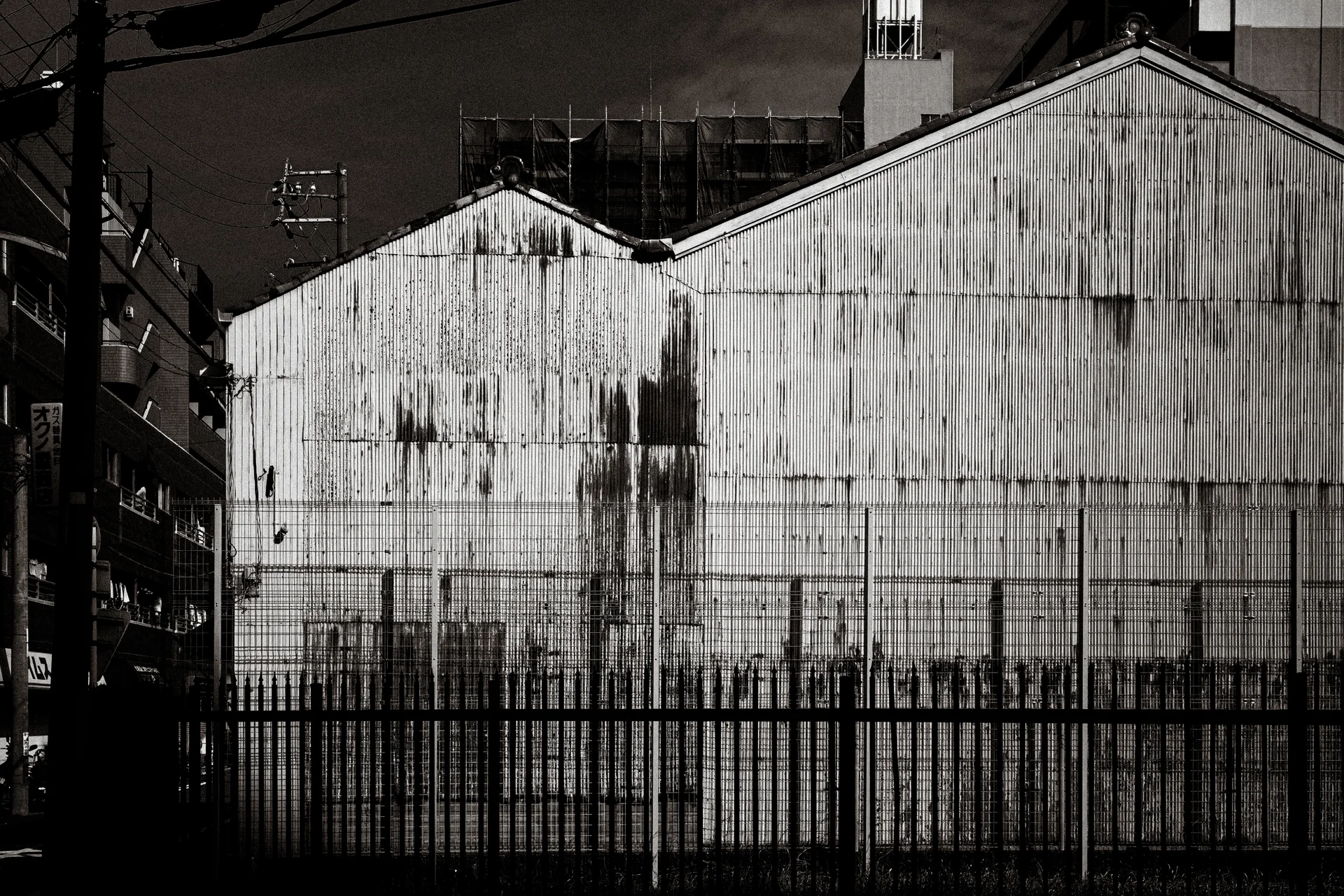 A stained corrugated warehouse wall with twin peaked roofs rises behind metal fencing in black and white.