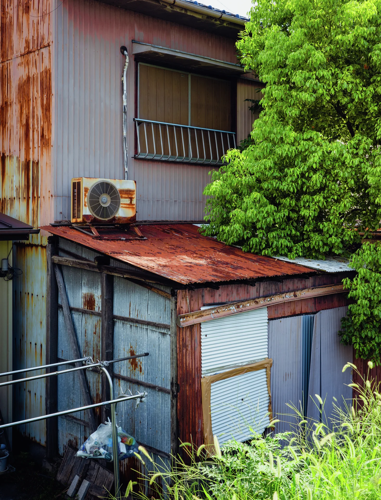 A rusty corrugated extension sits beneath an upstairs window and air conditioner beside dense green foliage.