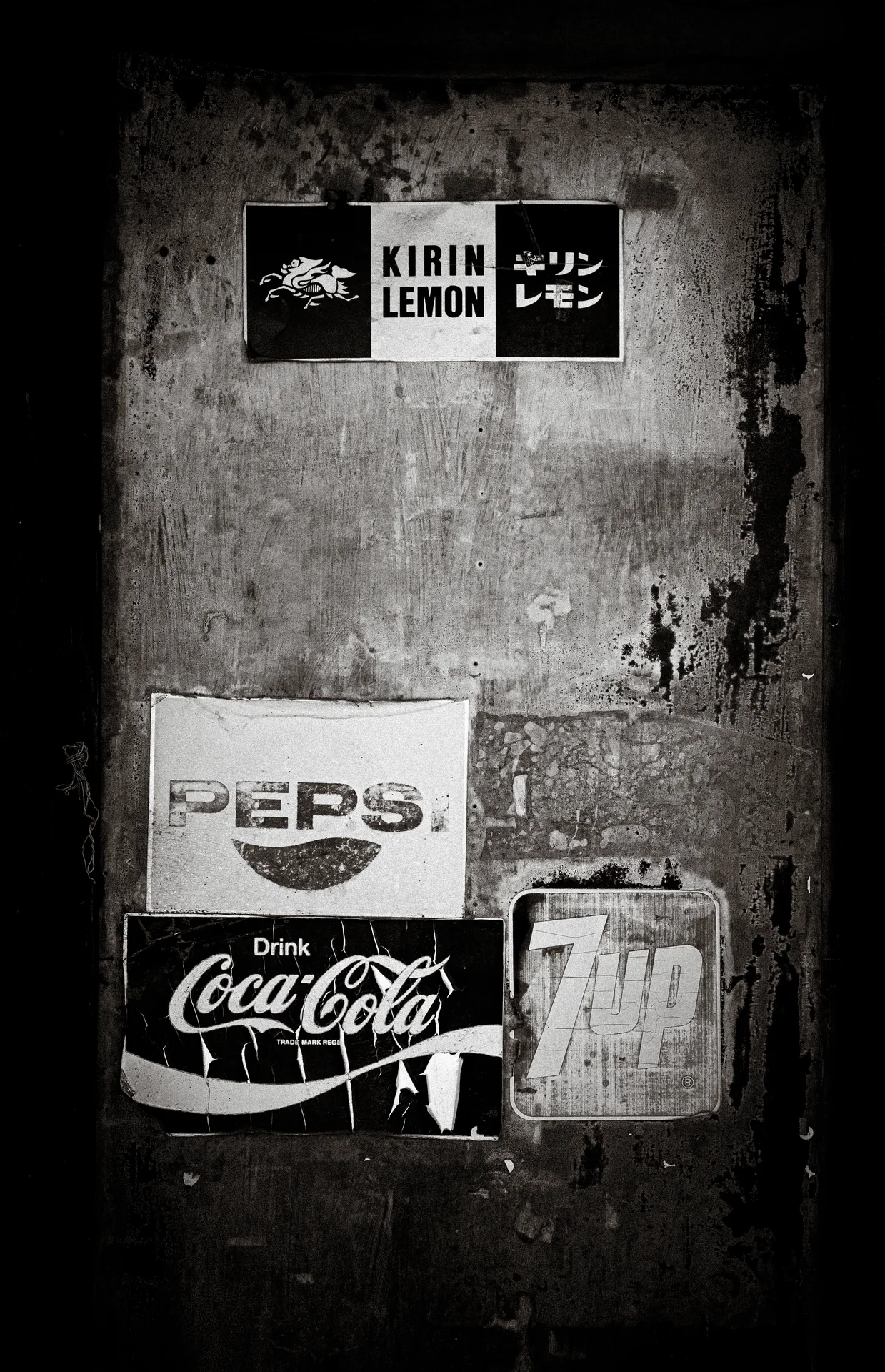 Old soft-drink advertisements cover a stained wall beside a small metal box in monochrome.