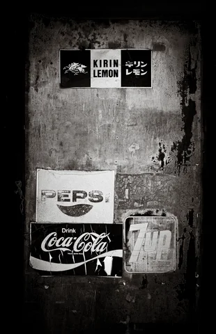 Old soft-drink advertisements cover a stained wall beside a small metal box in monochrome.
