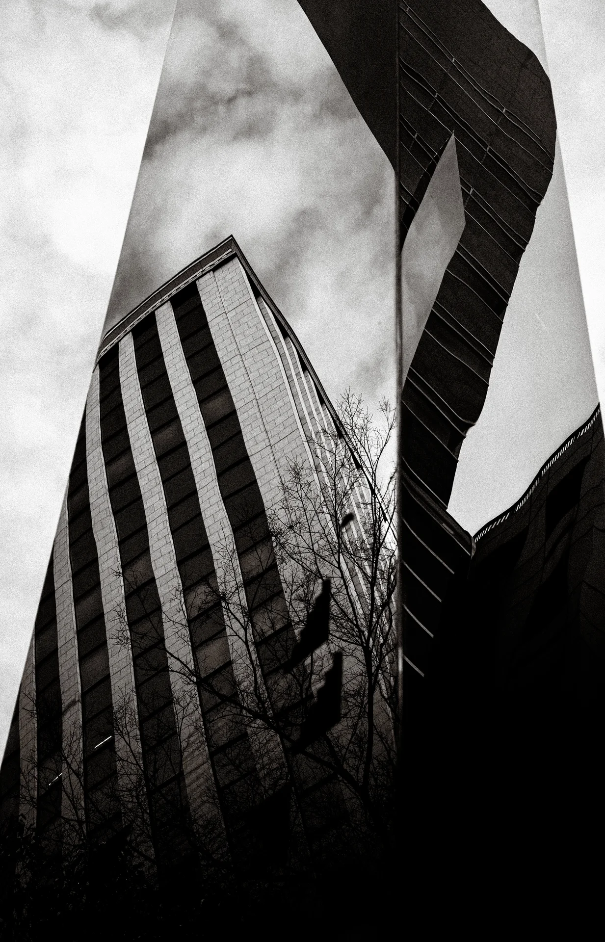Tall buildings rise at steep angles beside a dark projecting facade beneath clouds in black and white.