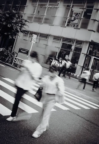 Blurred pedestrians cross white road stripes before a glass-fronted building in a tilted black and white view.