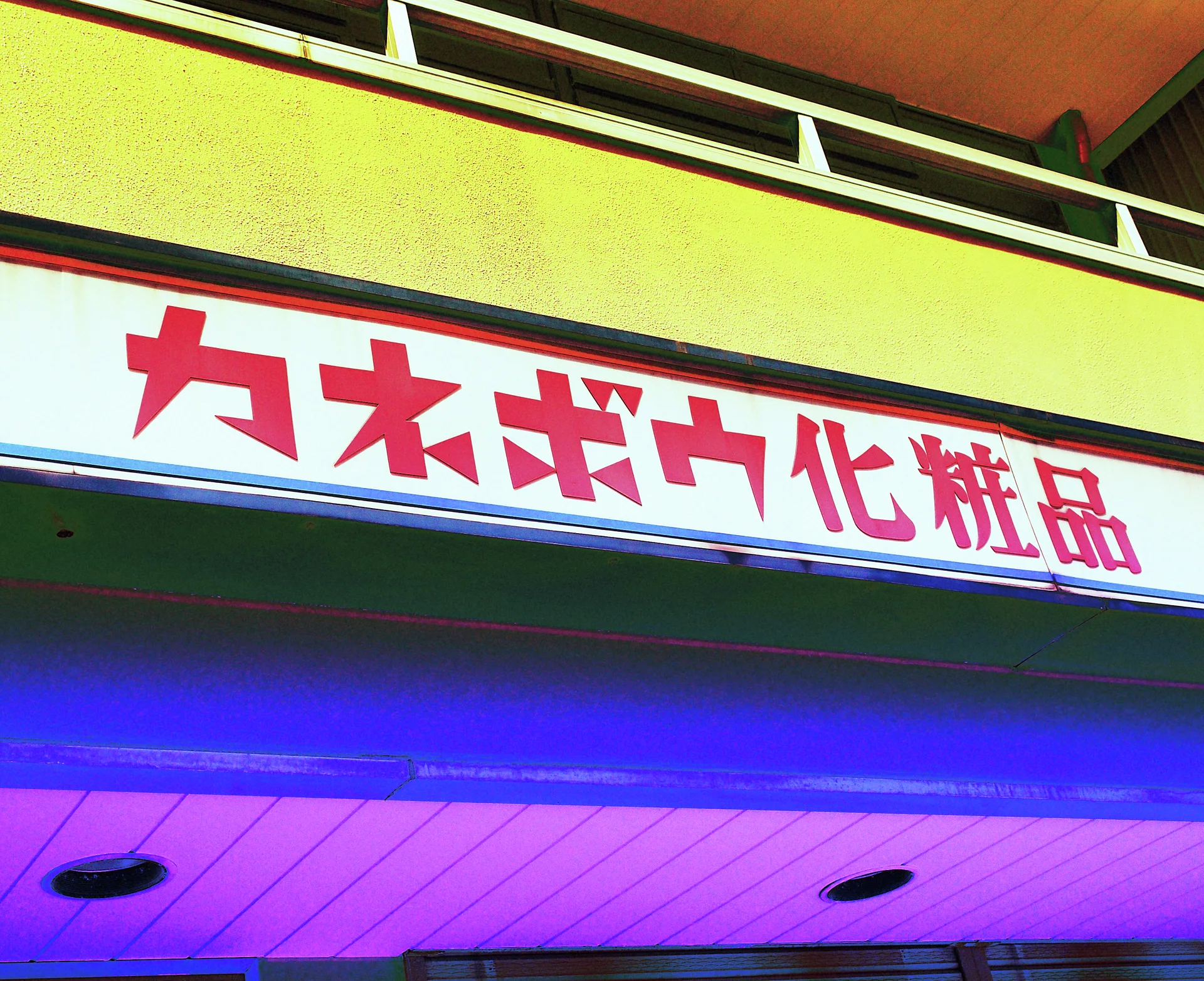 Bold red Japanese lettering spans a white sign beneath a yellow facade and above a purple-lit soffit.