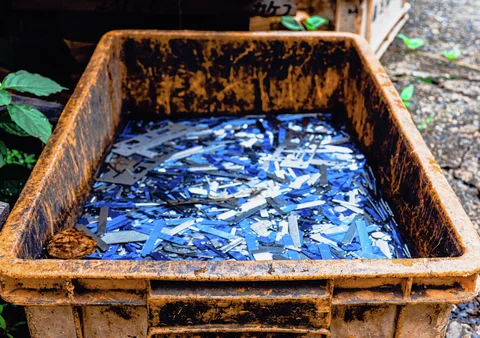 Blue and white scraps fill a large rust-stained metal bin standing beside gravel and green leaves.