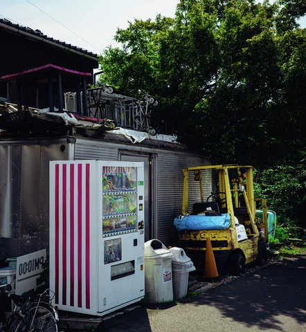 A striped drinks vending machine stands beside a yellow forklift beneath trees and a low corrugated shed.