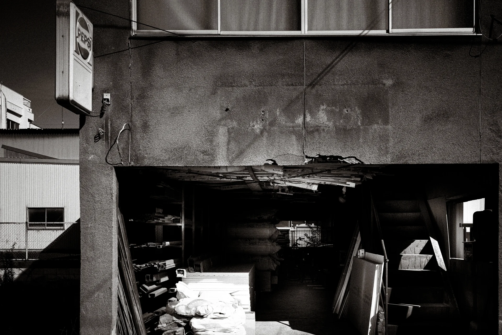 A damaged wall opening reveals stacked materials beneath cracked plaster and exposed timber in black and white.