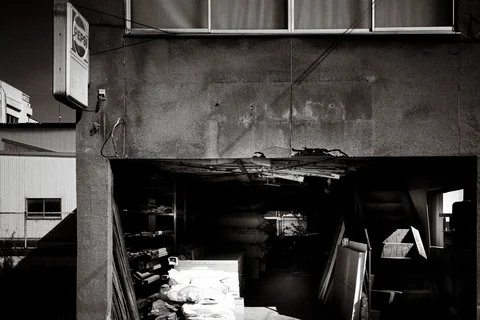 A damaged wall opening reveals stacked materials beneath cracked plaster and exposed timber in black and white.