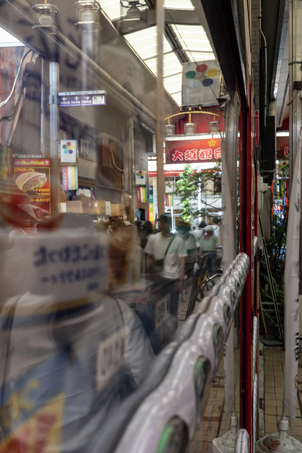 Layered window reflections show signs and pedestrians along a narrow passage beneath overhead beams.