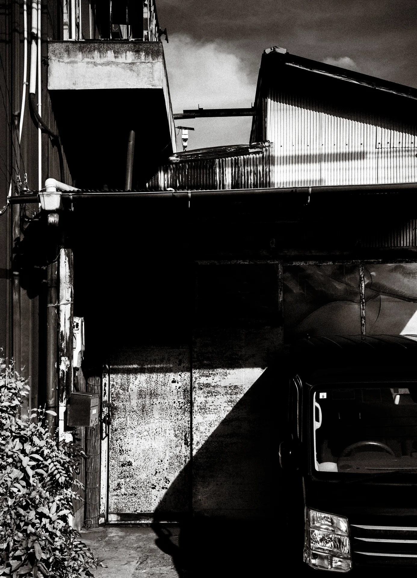 Sharp shadows divide a cramped gap between weathered buildings and a parked car in black and white.