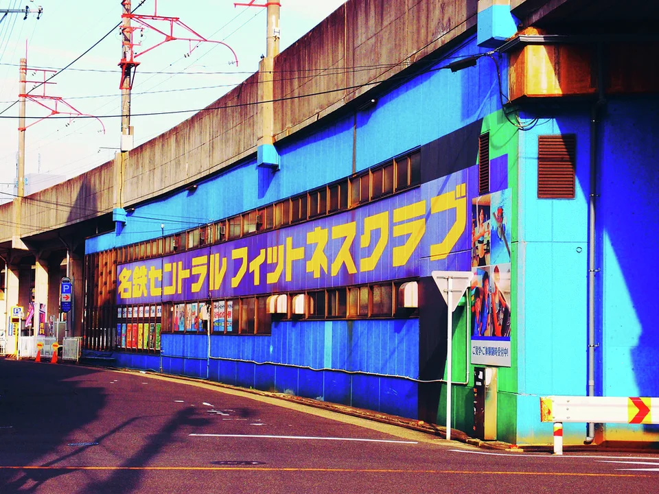 Large yellow Japanese lettering spans a bright blue building beneath an elevated concrete railway structure.