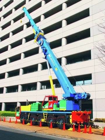 A blue mobile crane extends upward beside a multistory parking structure surrounded by orange traffic cones.