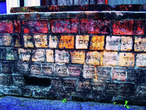 Weathered multicolored bricks with stamped lettering form a low wall above small weeds.