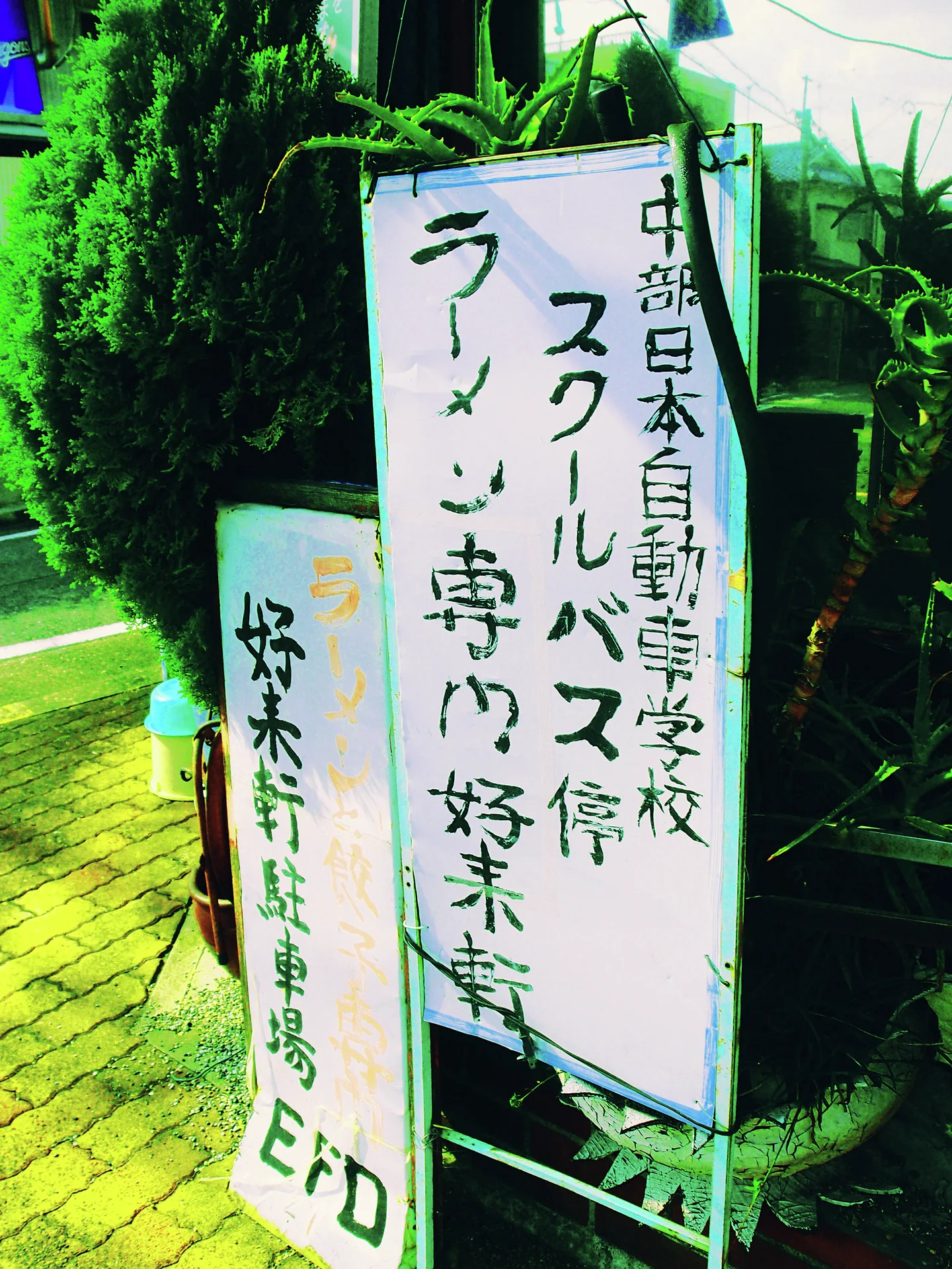 White boards bearing handwritten Japanese lettering stand beside a leafy sidewalk and dense greenery.