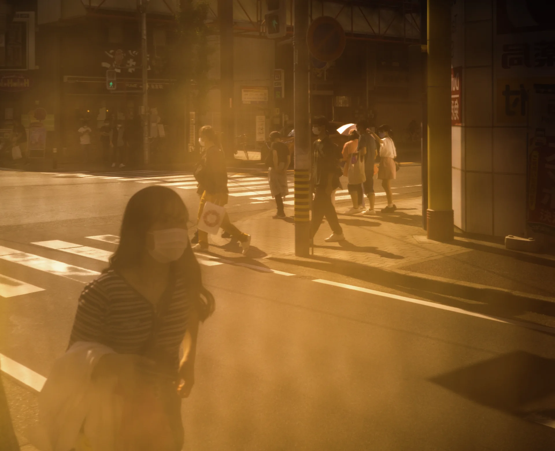 A woman stands near a sunlit crossing while pedestrians and their long shadows extend behind her.