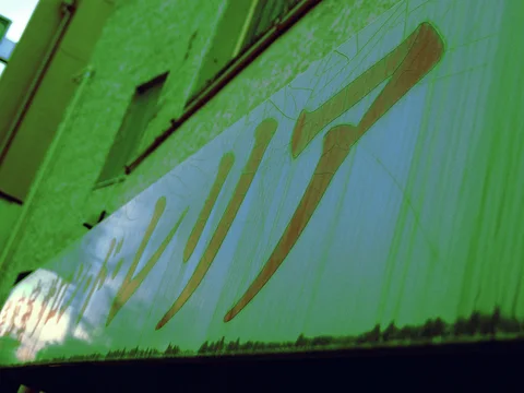 Faded gold characters stretch across a pale rust-streaked sign beneath a green wall.