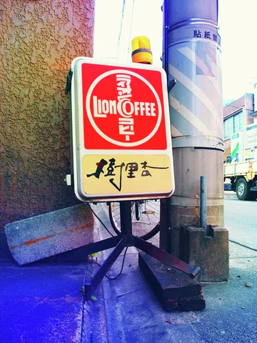 A red and cream coffee sign topped by a yellow light stands beside a striped utility pole.