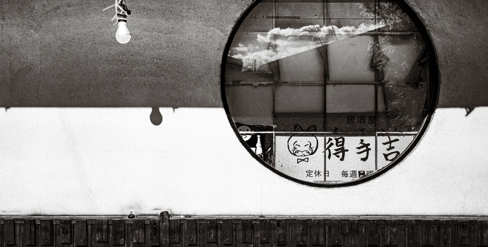 A round mirror reflects clouds and a sign above a white wall in black and white.