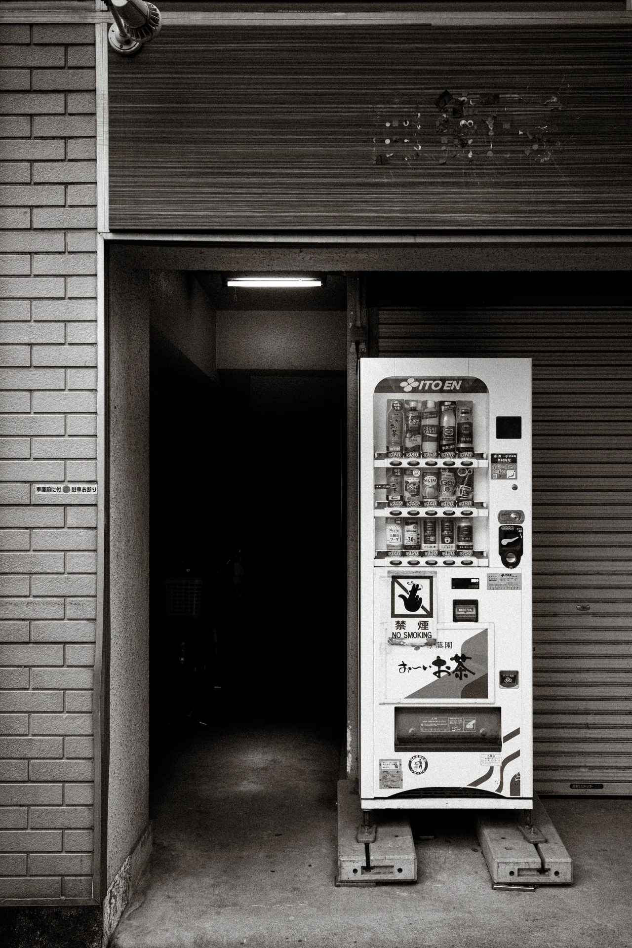 A narrow vending machine stands inside a recessed entrance beneath a shutter in monochrome.