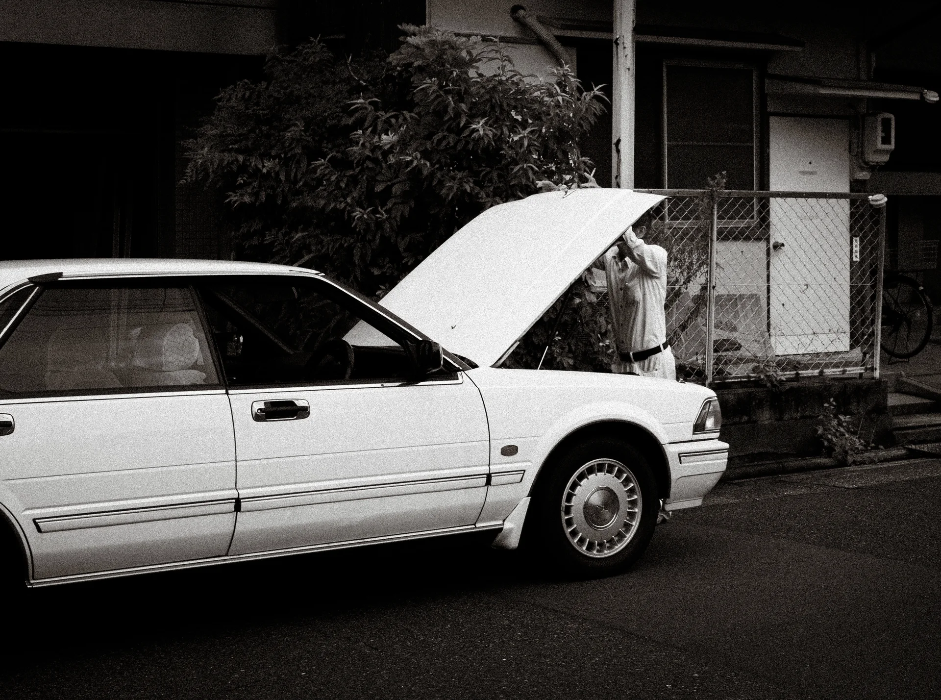 A white sedan has its hood raised beside a fenced building in black and white.