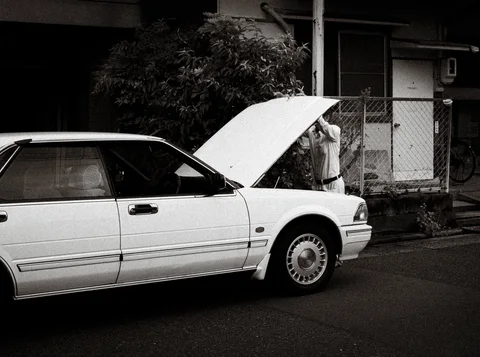 A white sedan has its hood raised beside a fenced building in black and white.