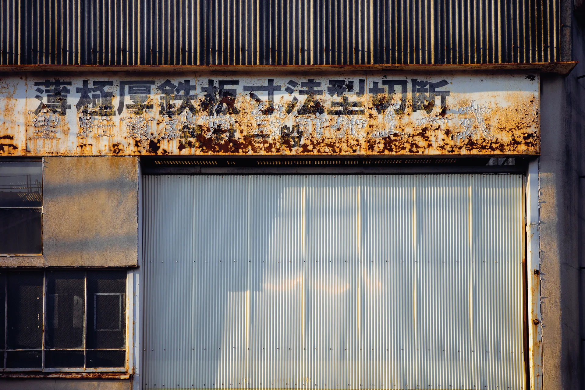 A rust-stained sign with faded lettering spans a corrugated shutter above a small dark window.