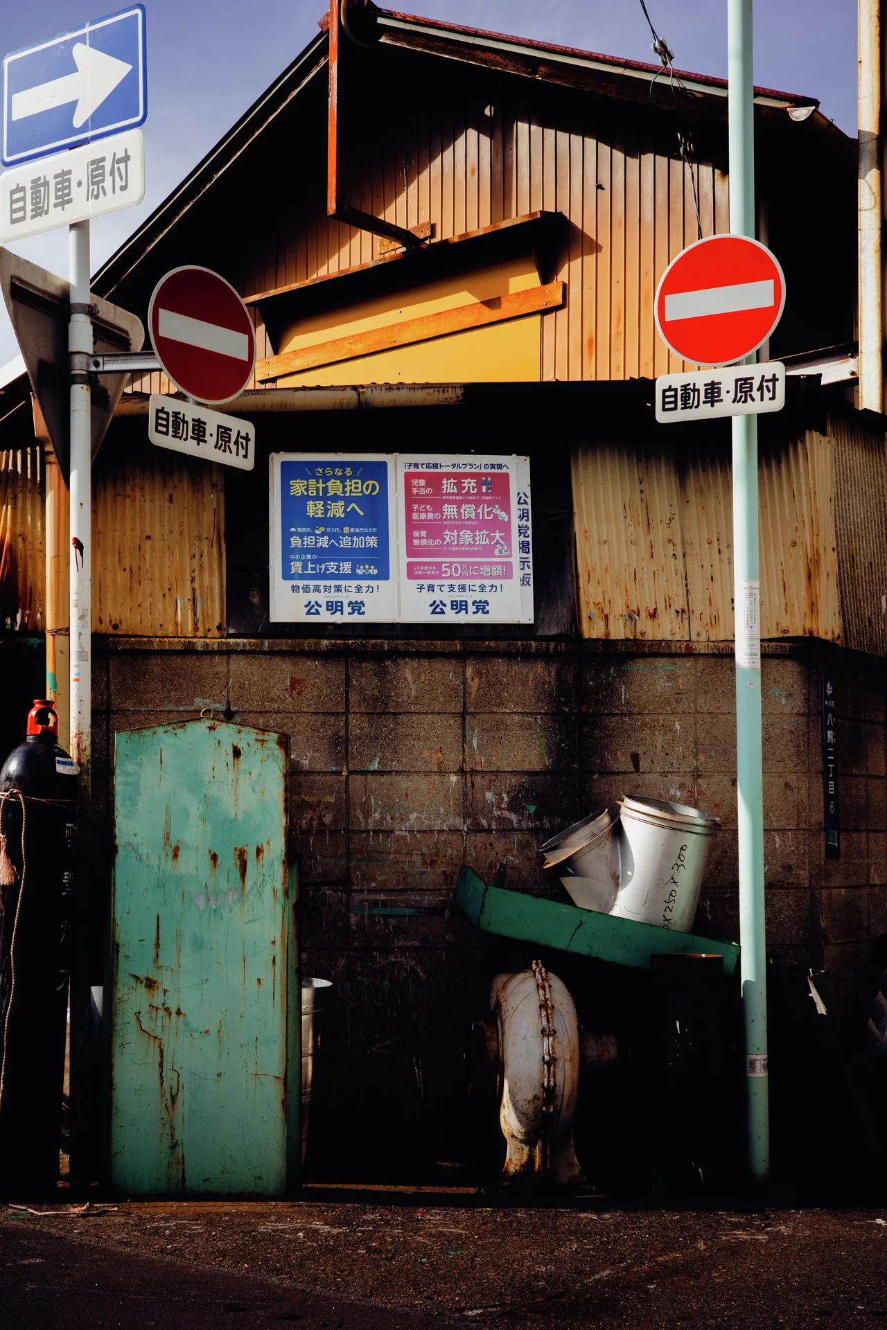 No-entry signs flank a wooden corner building with yellow trim and weathered turquoise panels.