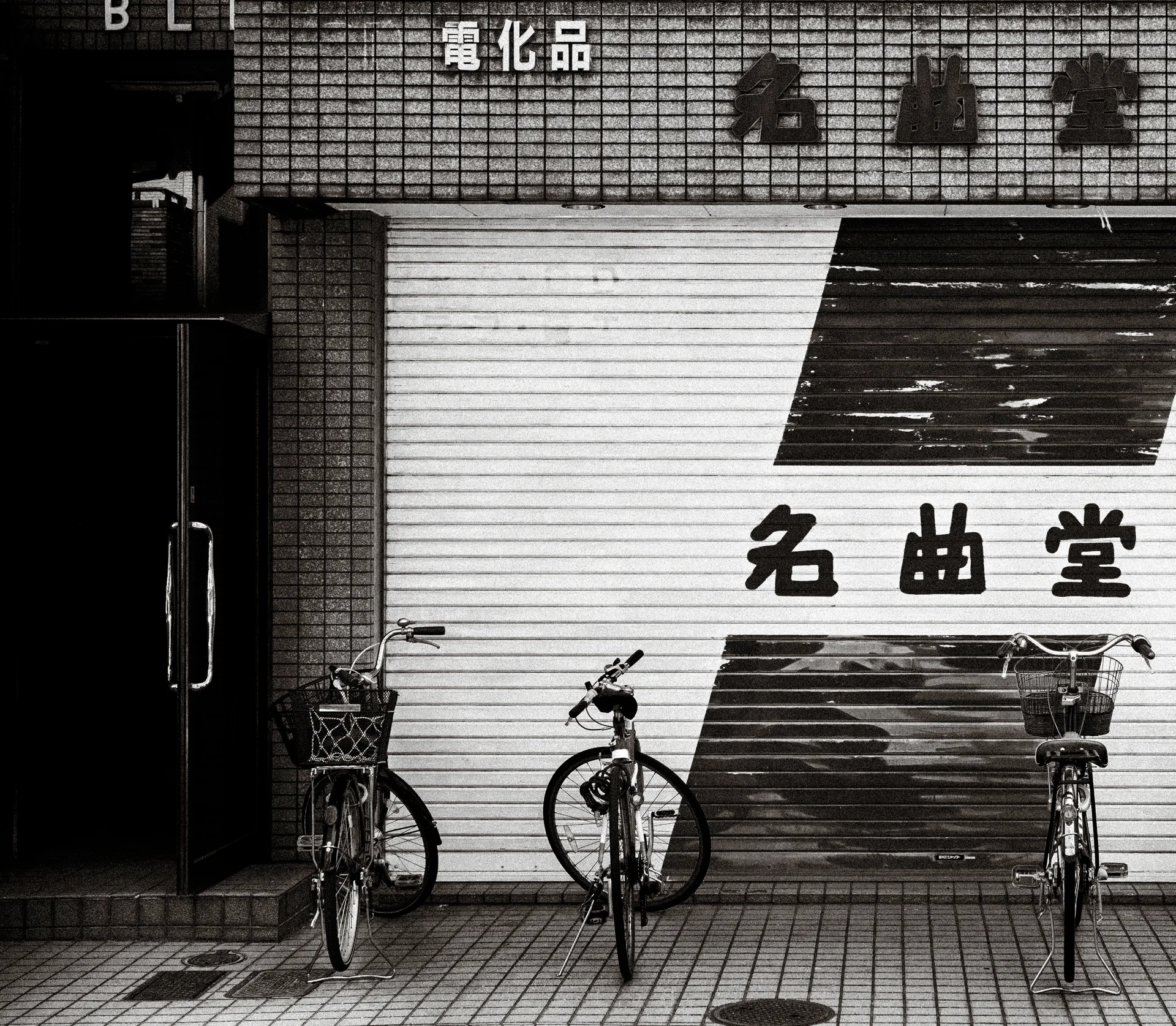 Three bicycles stand before a striped metal shop shutter with bold lettering in black and white.