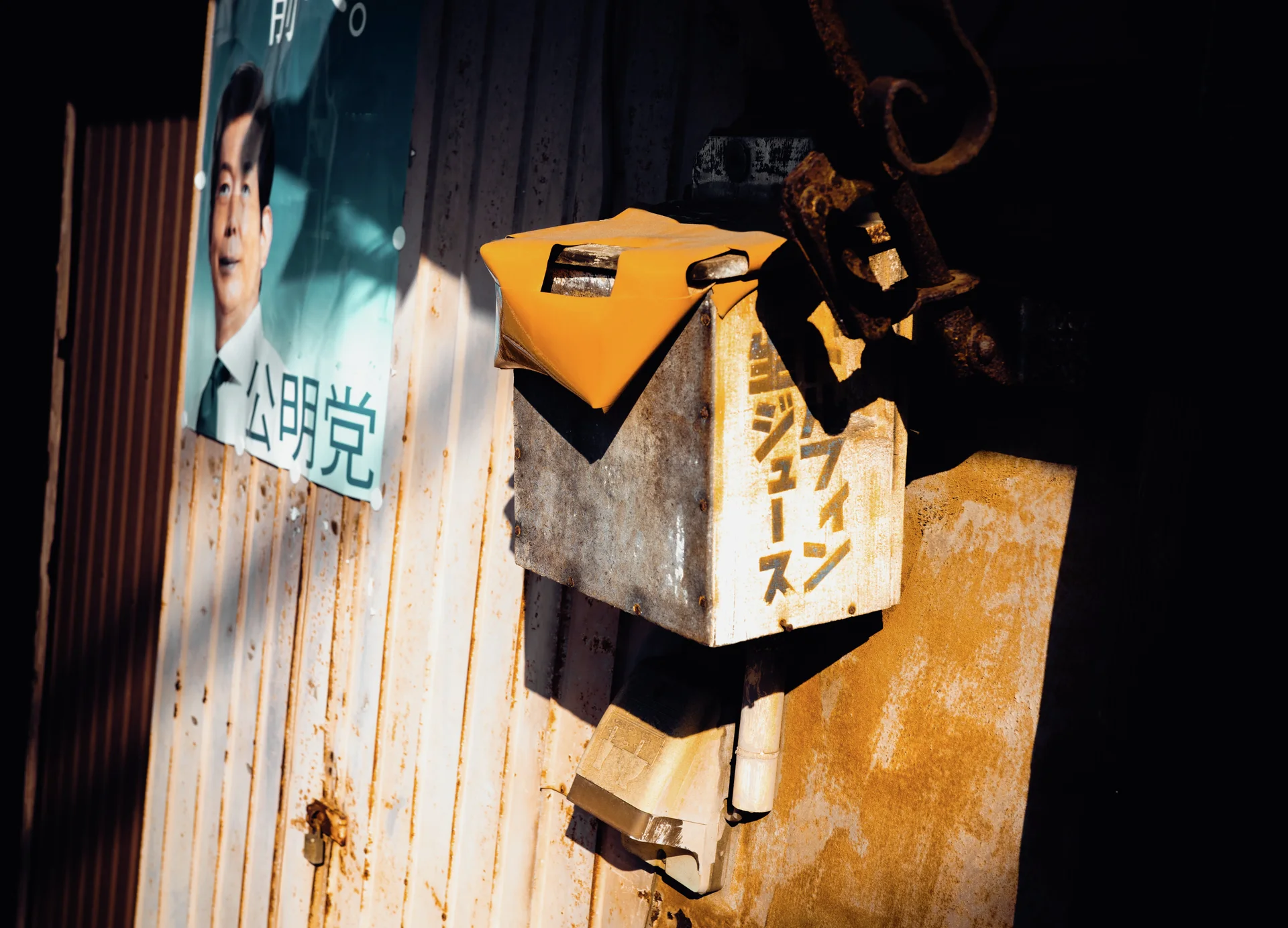 A yellow-topped metal box projects from weathered siding beside a portrait poster.