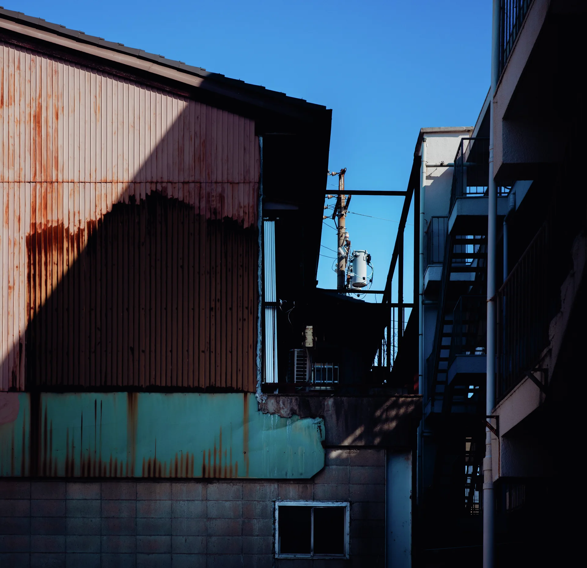 Deep triangular shadows fall across rust-streaked corrugated siding beside a narrow gap beneath blue sky.