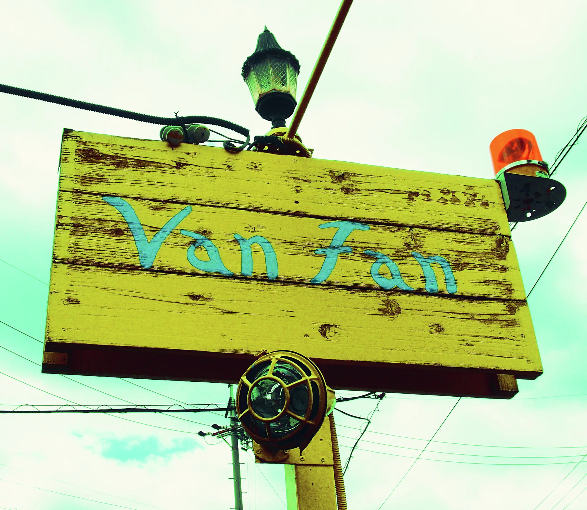 Blue Van Fan lettering spans a weathered yellow wooden sign beneath lamps and cables.