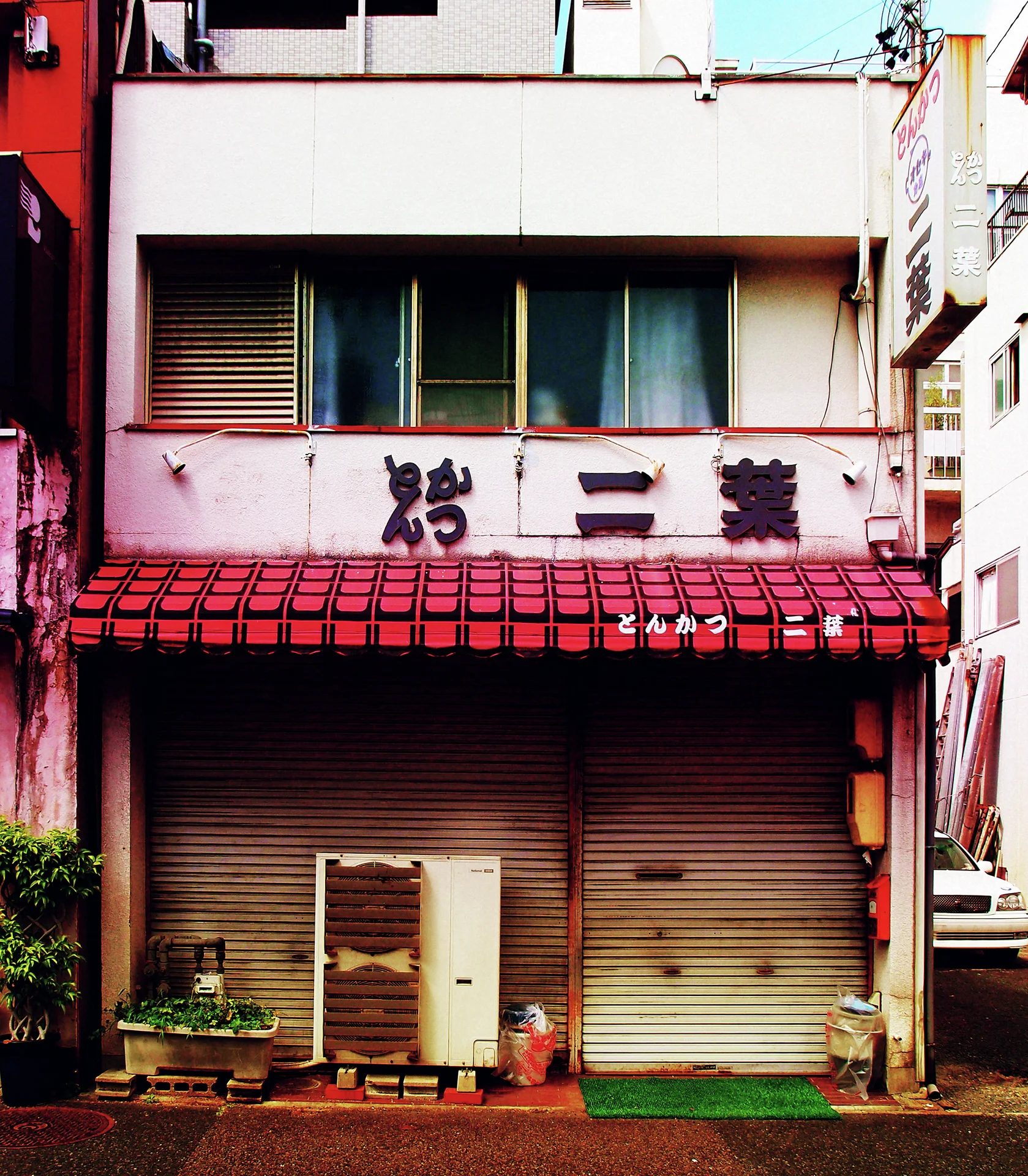 A red tiled awning shelters closed restaurant shutters beside an air conditioner and small pots.