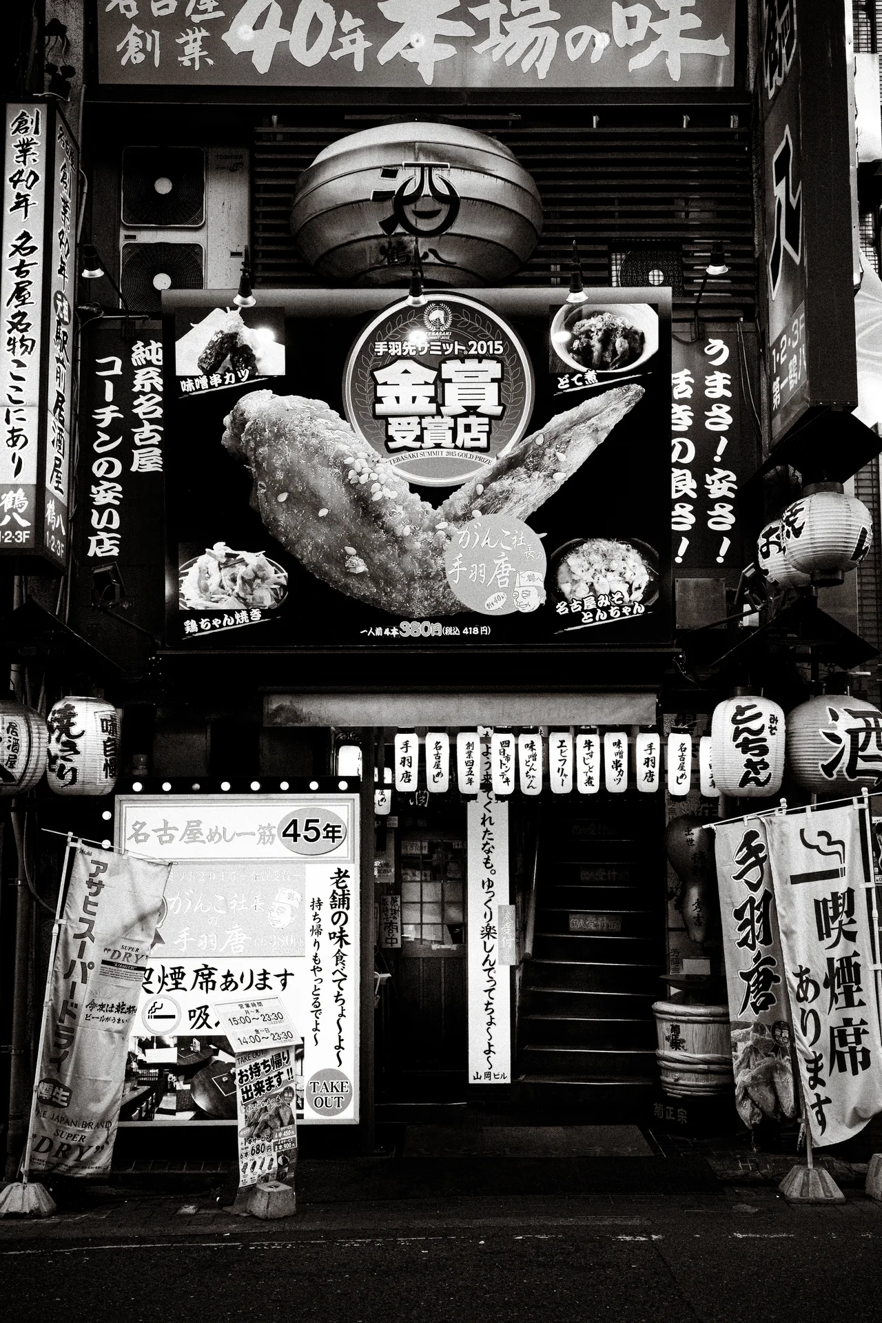 Lanterns and densely arranged illustrated signs surround a narrow restaurant entrance in a black and white view.