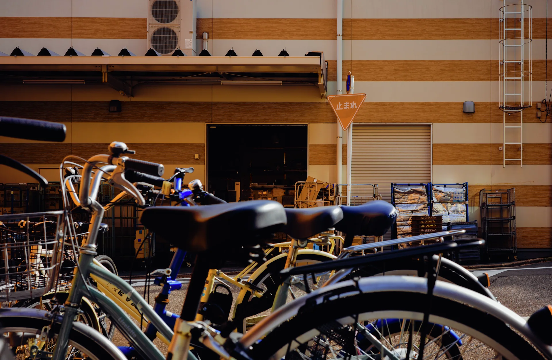 Bicycle saddles and handlebars fill the foreground before a building painted in broad yellow and white stripes.