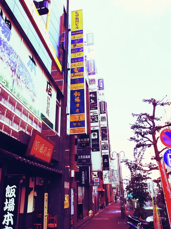 Multiple tall colorful shop directories rise above a narrow sidewalk lined with buildings and closely pruned trees.