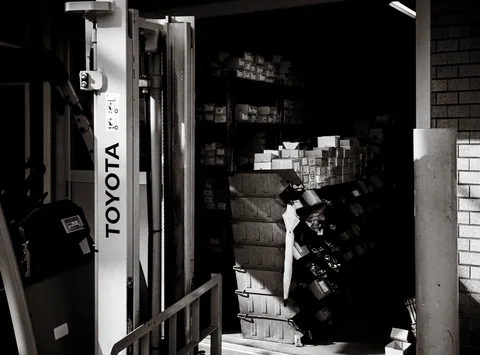 Stacks of boxes fill a dark workshop beside a white Toyota-marked post in black and white.