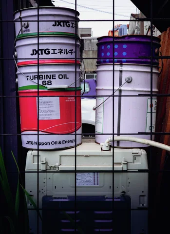 Stacked oil drums sit behind dark metal bars beside industrial equipment.