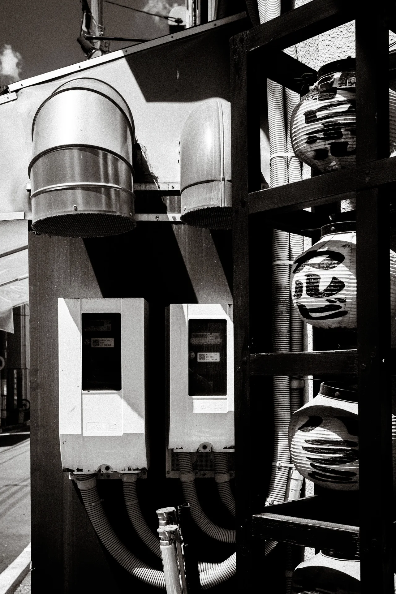 Metal ventilation ducts, utility boxes, and stacked lanterns crowd an exterior wall in monochrome.