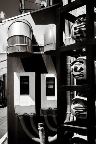 Metal ventilation ducts, utility boxes, and stacked lanterns crowd an exterior wall in monochrome.