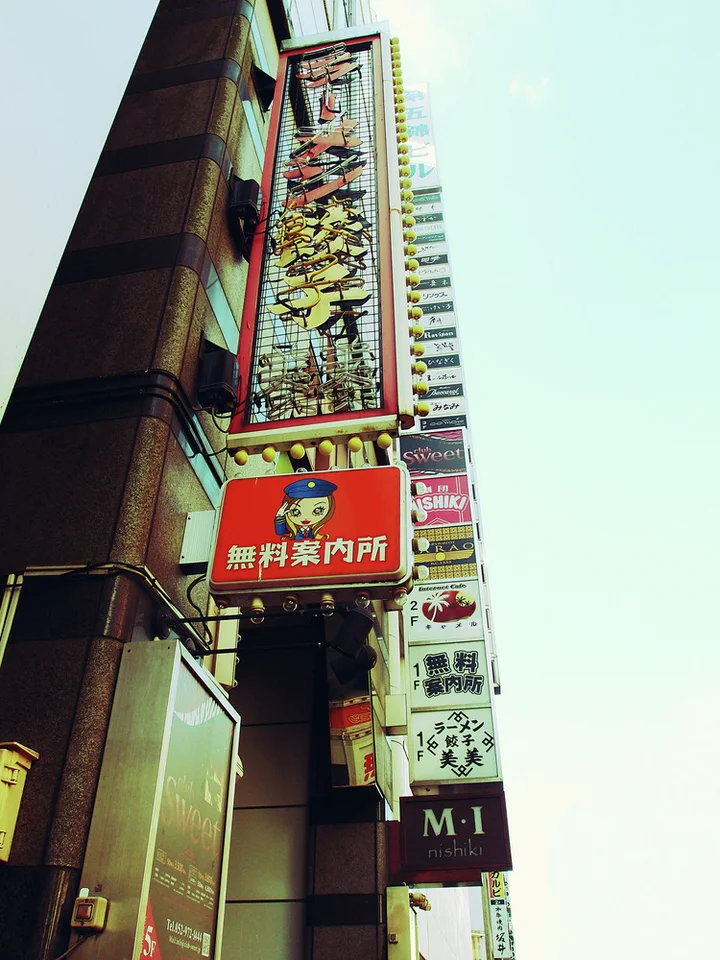 Dense vertical shop signs climb a narrow building facade above a red sign and exposed pipes.