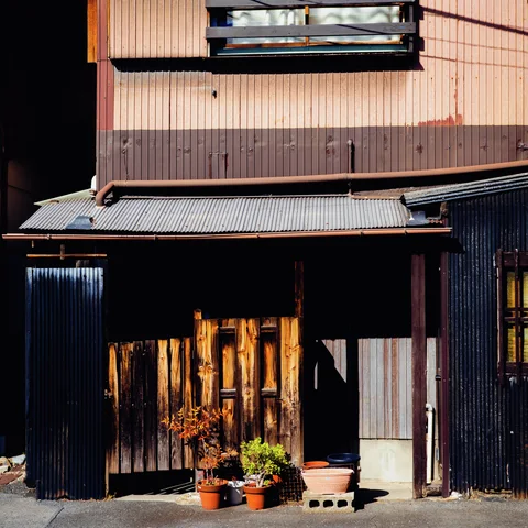 Potted plants stand outside a shadowed wooden entrance beneath a corrugated awning and faded pink upper wall.