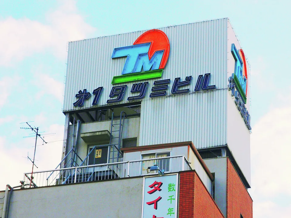 A colorful rooftop emblem and raised Japanese lettering mark a pale corrugated building beneath blue sky.
