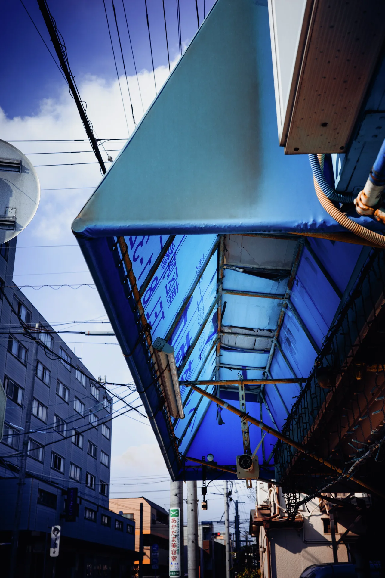 A translucent blue awning projects over a sidewalk beneath overhead wires and pale clouds.