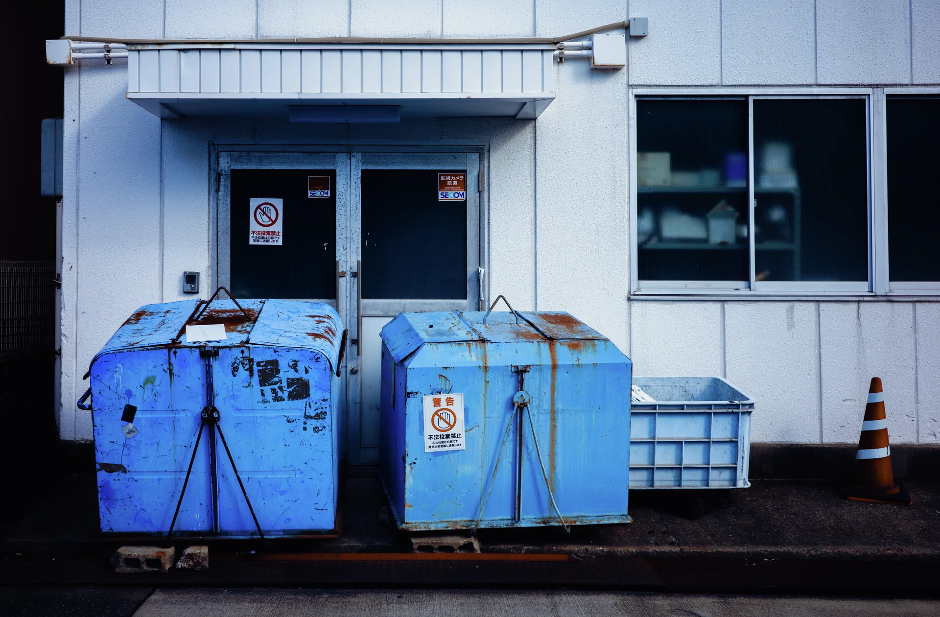 Two battered blue metal containers stand outside pale double doors beside a smaller bin and traffic cone.
