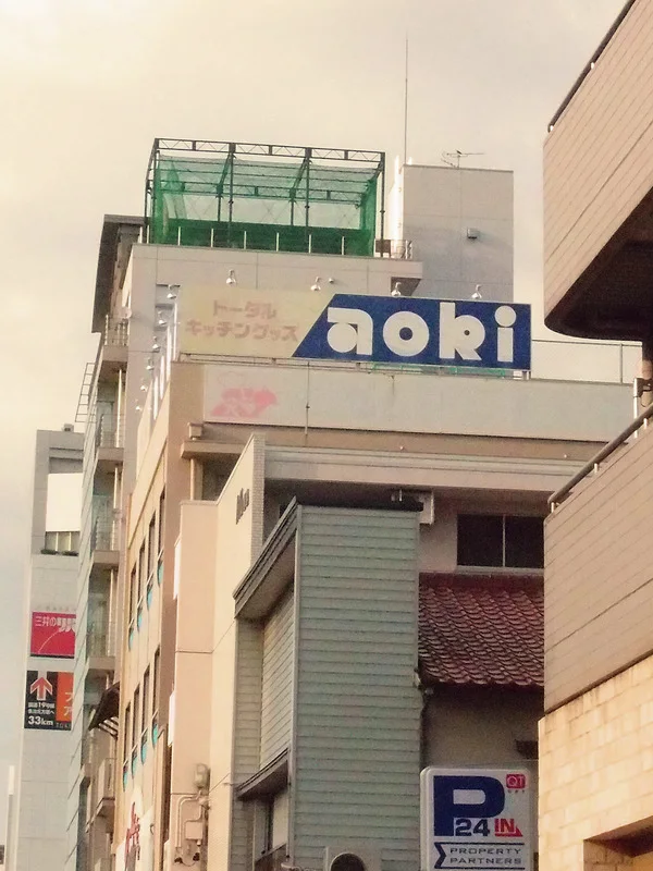 A blue Aori sign sits high on a pale building with a green rooftop enclosure.