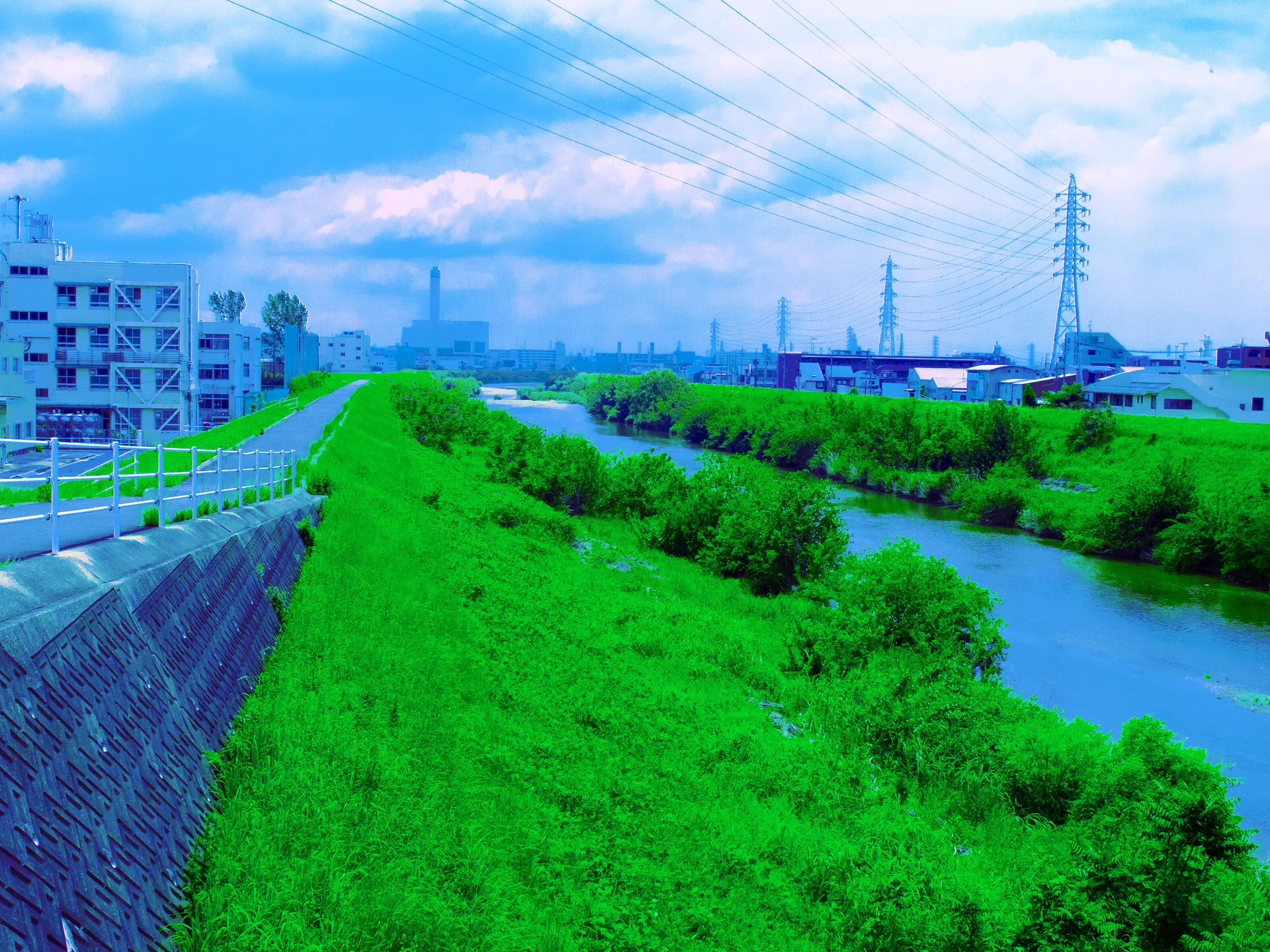 A river curves beside a broad grassy embankment beneath power lines, distant buildings, and scattered clouds.