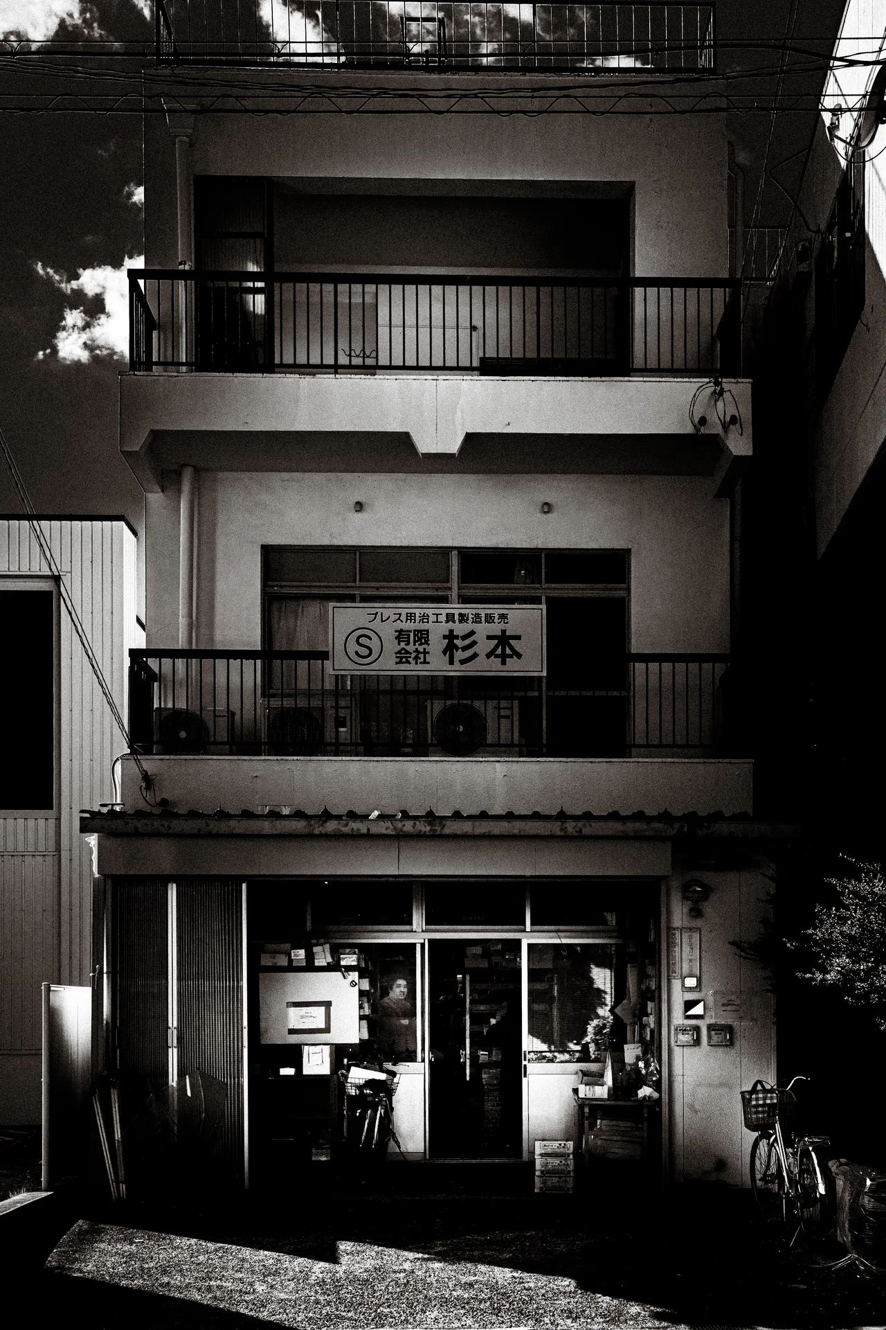 Stacked balconies rise above a glass-fronted entrance with a bicycle nearby in black and white.