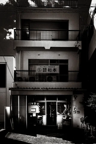 Stacked balconies rise above a glass-fronted entrance with a bicycle nearby in black and white.