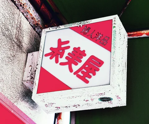 Red Japanese lettering and corner triangles fill a square projecting sign with a chipped white frame.