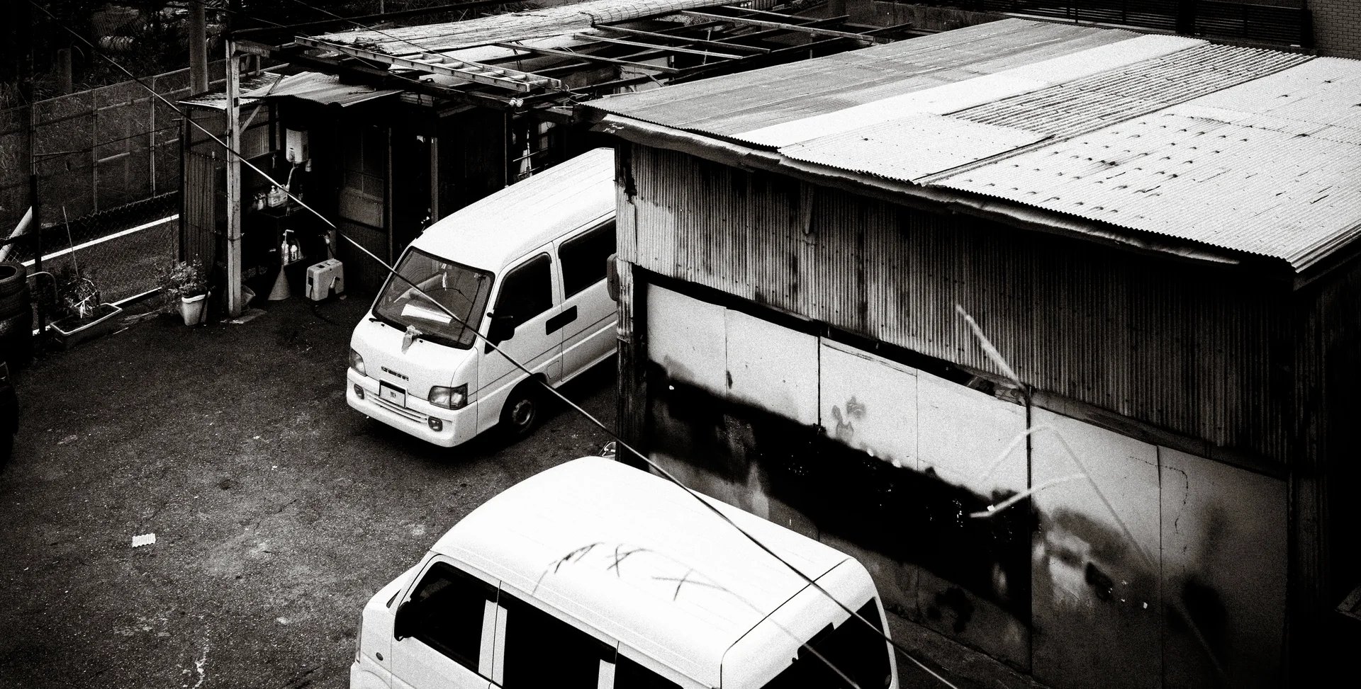 Two white vans occupy a gravel yard beside a weathered corrugated shed in black and white.