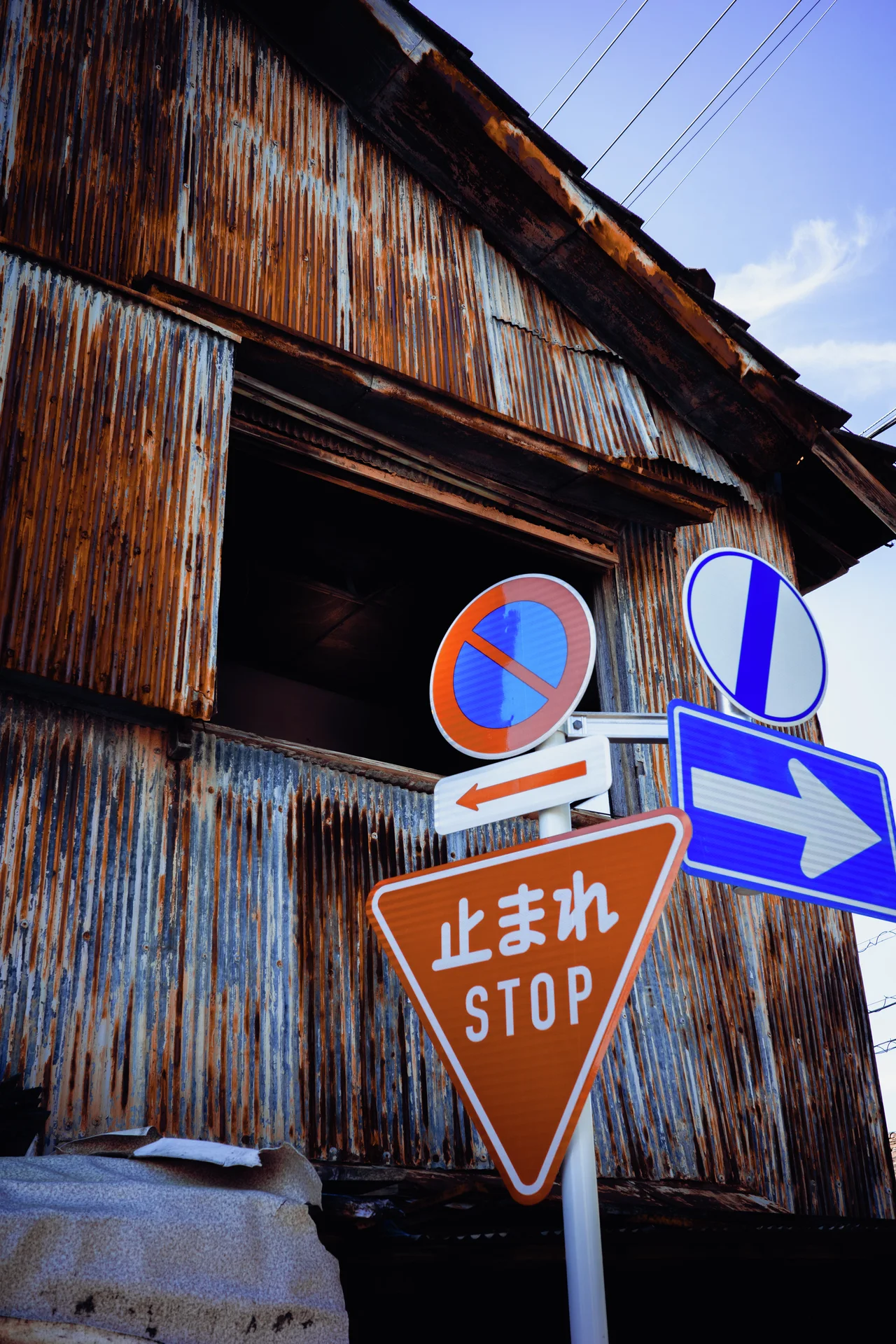 A stop sign and blue directional arrow stand before a rust-streaked corrugated building.